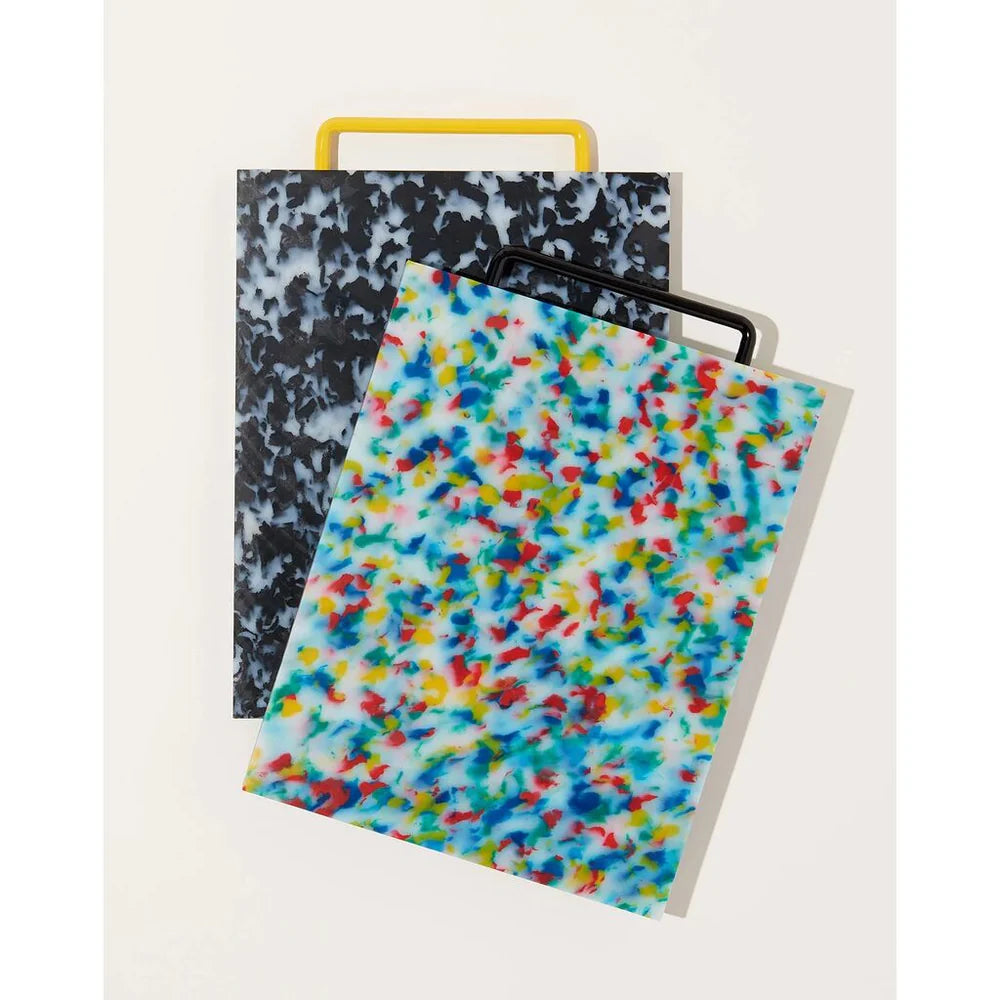 Two cutting boards one black and white confetti with a yellow handle the other red blue yellow and white confetti with a black handle.