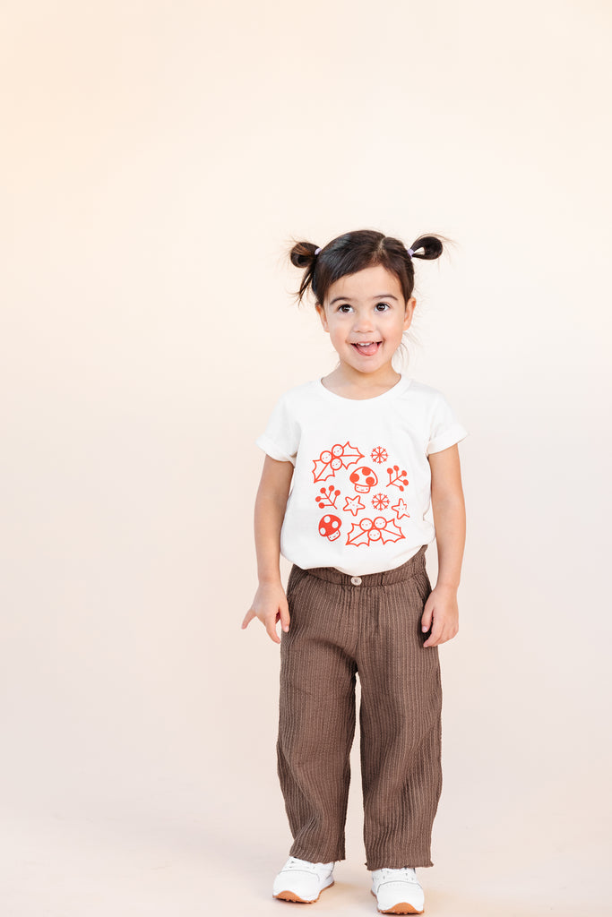 A child modeling a white tee screen printed with smiling mistletoe, snowflakes, mushrooms, leaves, and stars in red.