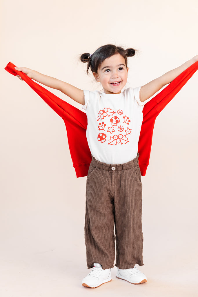 A child modeling a white tee screen printed with smiling mistletoe, snowflakes, mushrooms, leaves, and stars in red.