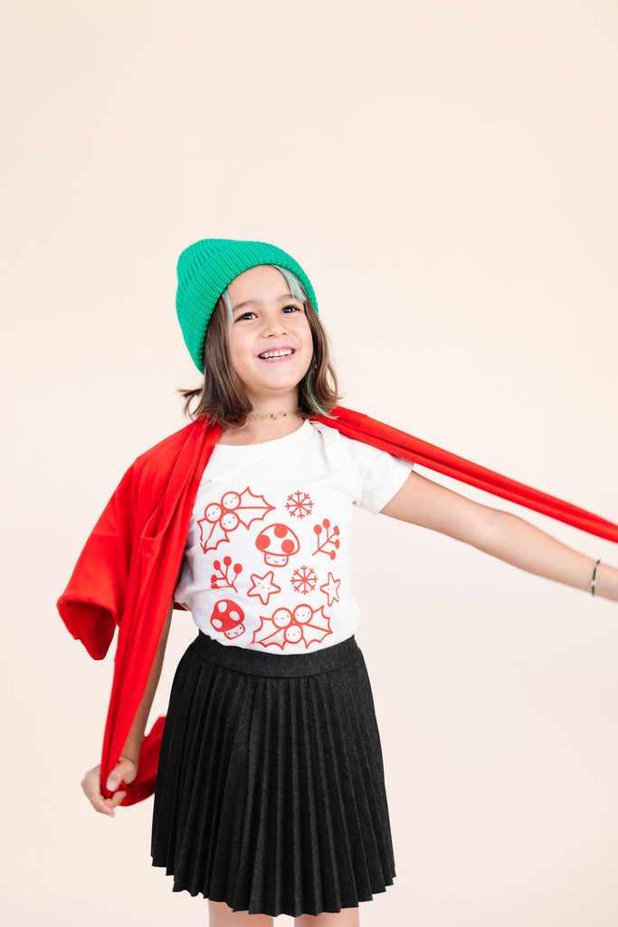A child modeling a white tee screen printed with smiling mistletoe, snowflakes, mushrooms, leaves, and stars in red.
