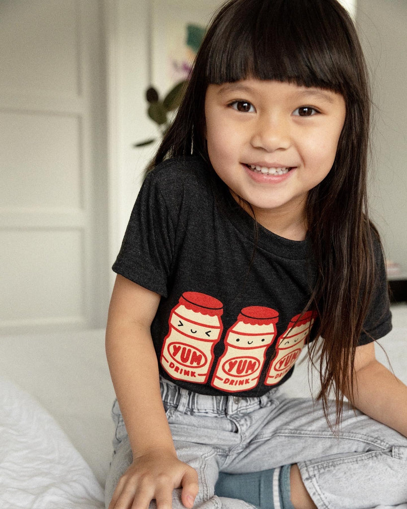 A child modeling a black tee screen printed with three red and yellow yakult yogurt bottles with smiley faces.