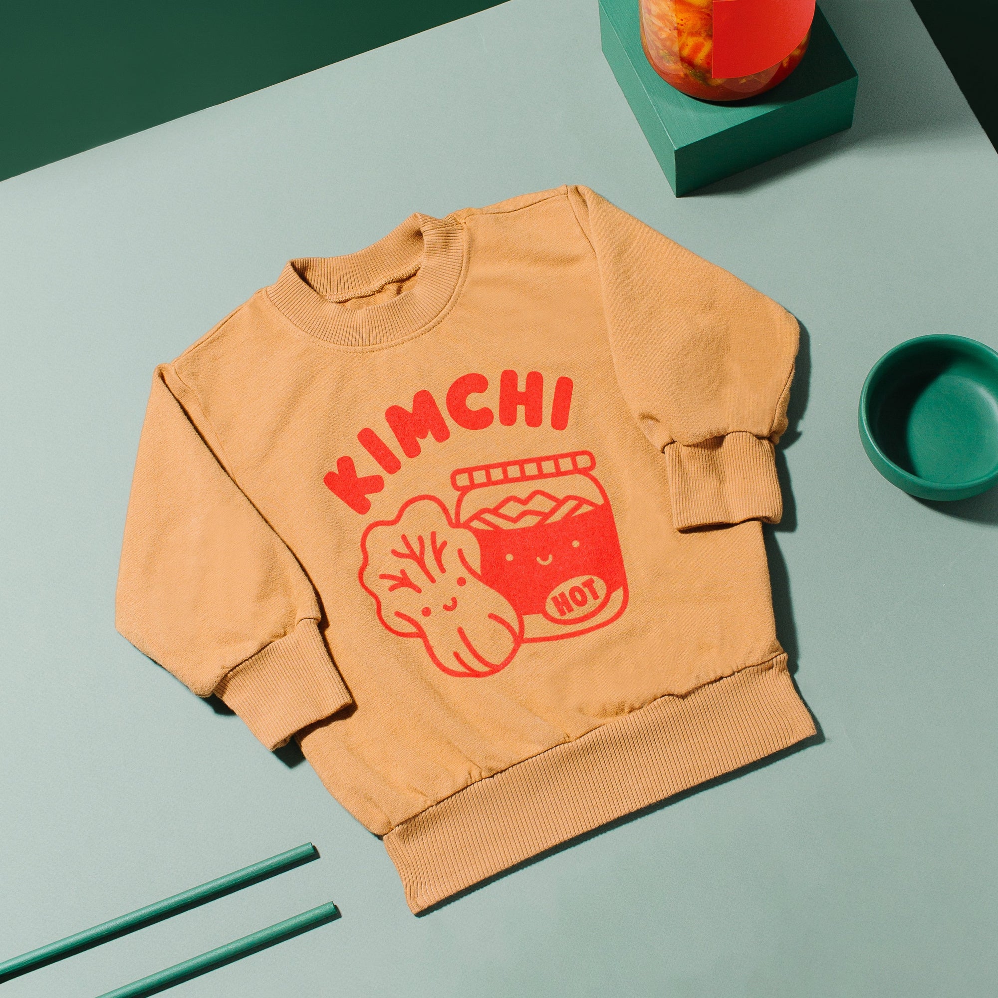 Brown sweatshirt screen printed with red ink, text KIMCHI and cabbage and jar of kimchi, both smiling.