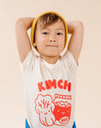 A child modeling a white tee screen printed with red ink, text KIMCHI and cabbage and jar of kimchi, both smiling.