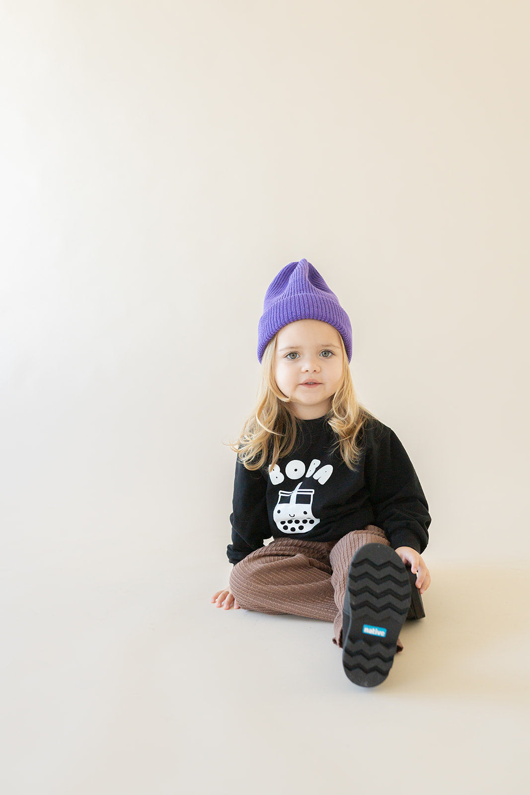 A child modeling a black sweatshirt with screen printed white ink text BOBA above a smiling glass of boba.