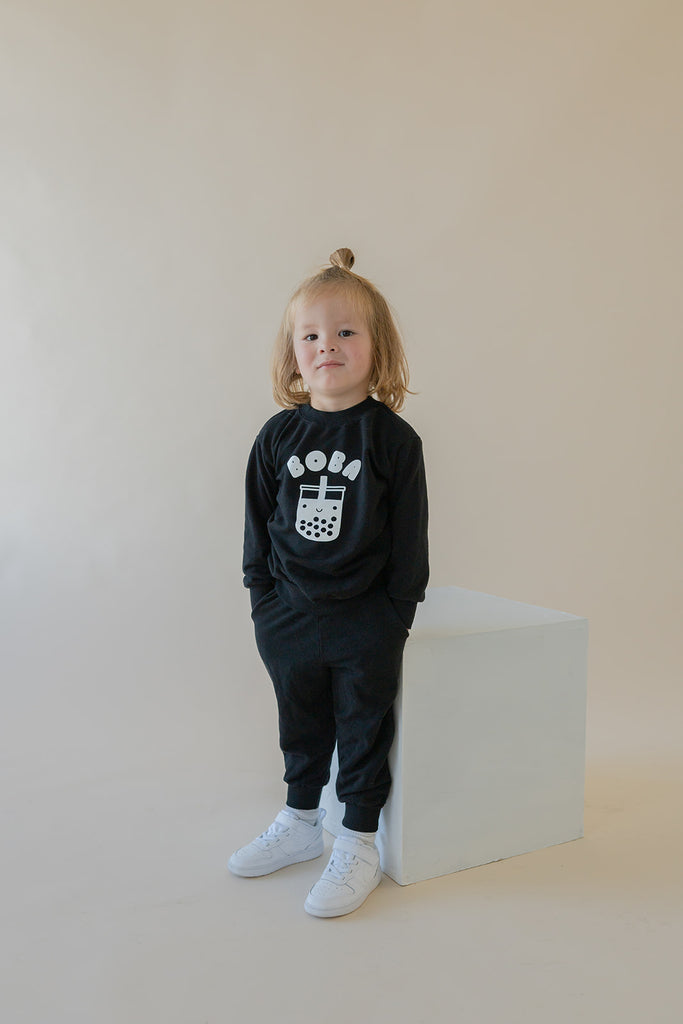 A child modeling a black sweatshirt with screen printed white ink text BOBA above a smiling glass of boba.